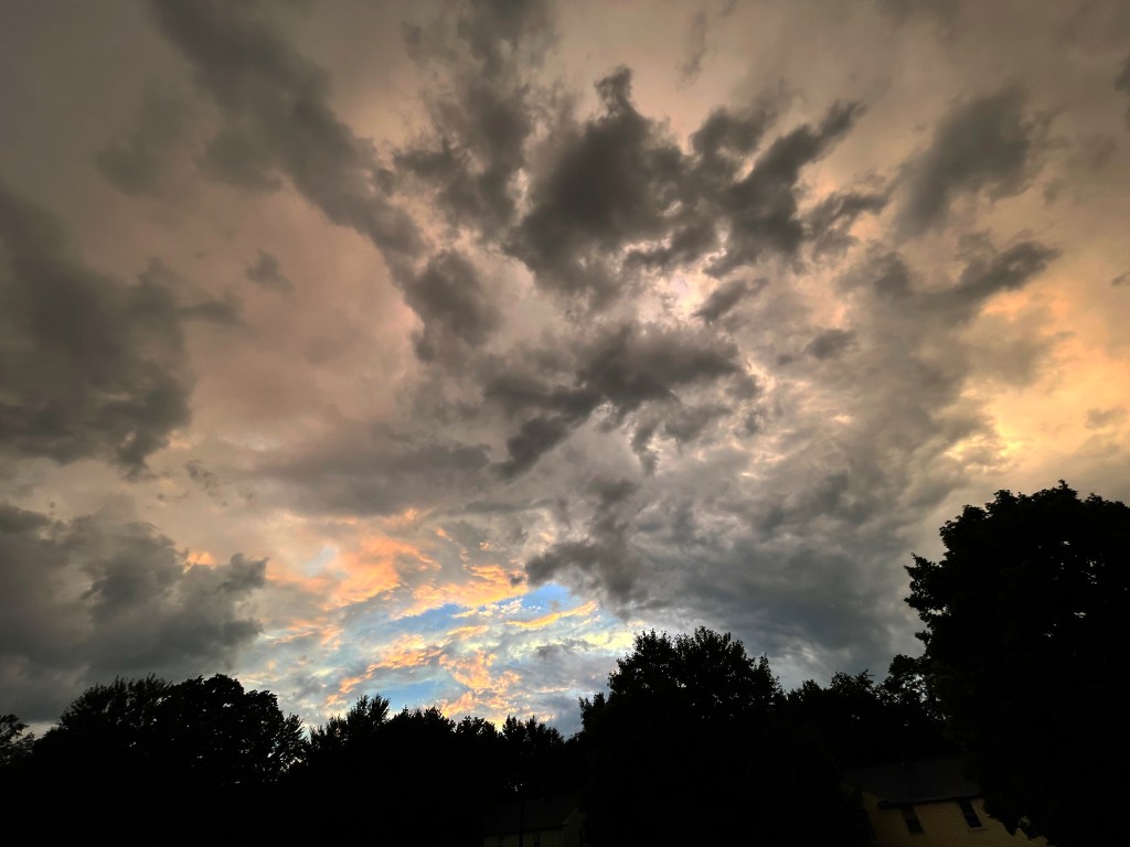 "Sunset After the Storm," photograph of luminously dark clouds scattering in a bright blue and golden sky after a rainstorm, with dark trees below